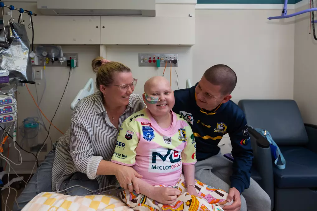 Emma sitting on hospital bed with her mum and dad. She is wearing the jersey she designed for Lakes United Rugby League Charity day.